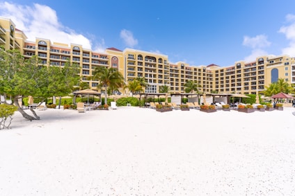 A beachfront resort with a large, elegant building featuring multiple stories and balconies, surrounded by palm trees. The foreground consists of a white sandy beach with scattered lounge chairs and umbrellas. The sky is clear with a few clouds, indicating a sunny day.