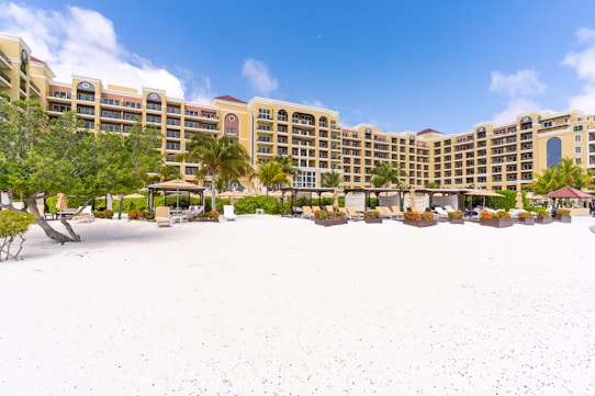 A beachfront resort with a large, elegant building featuring multiple stories and balconies, surrounded by palm trees. The foreground consists of a white sandy beach with scattered lounge chairs and umbrellas. The sky is clear with a few clouds, indicating a sunny day.