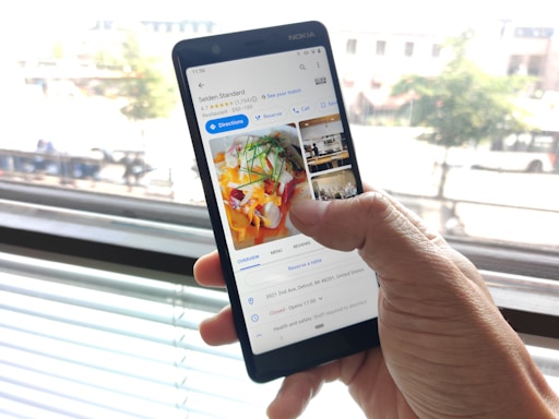 A hand is holding a smartphone in front of a window. On the screen, a Google search for 'Selden Standard' is displayed, showing images and details of the restaurant including ratings and directions.