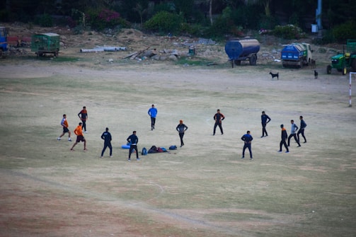 A group of eleven people engaged in an outdoor activity on an open dusty field. They are standing in a circle, possibly participating in a team exercise or discussing a strategy. Around them, there are scattered bags and equipment on the ground. The background shows a few parked vehicles and some greenery alongside the edge of the field.