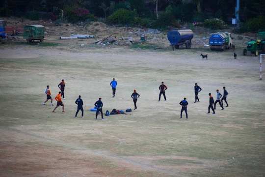 A group of eleven people engaged in an outdoor activity on an open dusty field. They are standing in a circle, possibly participating in a team exercise or discussing a strategy. Around them, there are scattered bags and equipment on the ground. The background shows a few parked vehicles and some greenery alongside the edge of the field.