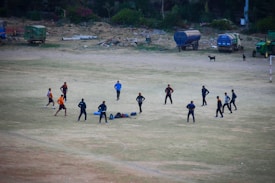 A group of eleven people engaged in an outdoor activity on an open dusty field. They are standing in a circle, possibly participating in a team exercise or discussing a strategy. Around them, there are scattered bags and equipment on the ground. The background shows a few parked vehicles and some greenery alongside the edge of the field.