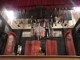 A theatrical stage setup with a red curtain and numerous costumes hanging from a chandelier. The backdrop includes abstract panels with Asian-inspired designs. To the sides are large, ornate wooden mirrors.