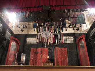 A theatrical stage setup with a red curtain and numerous costumes hanging from a chandelier. The backdrop includes abstract panels with Asian-inspired designs. To the sides are large, ornate wooden mirrors.