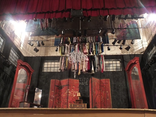 A theatrical stage setup with a red curtain and numerous costumes hanging from a chandelier. The backdrop includes abstract panels with Asian-inspired designs. To the sides are large, ornate wooden mirrors.