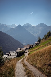 A serene mountain landscape with a winding dirt path leading through a small village of rustic houses. The path is flanked by green and golden grass fields, with majestic mountains towering in the background under a clear blue sky.