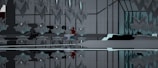 An abstract interior space with symmetrical geometric structures and mirrored surfaces. The design features sharp, angular elements with a modern aesthetic. A notable touch of red and blue hints at the presence of an abstract object or character nestled within the environment, creating a focal point amid the predominantly gray and black tones.