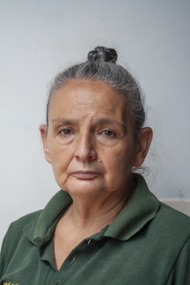 An older woman with grey hair pulled back in a bun is wearing a dark green collared shirt. Her expression is serious and pensive, and she is positioned against a plain, light-colored background.