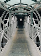 A long, narrow metal walkway extends forward, enclosed by a series of intersecting metal beams and a transparent, arched roof. The walkway is flanked by metal railings on both sides. The structure creates a tunnel-like effect with repeating patterns of geometric shapes, providing an industrial and symmetrical aesthetic.