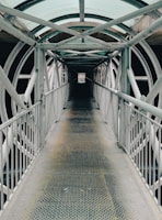 A long, narrow metal walkway extends forward, enclosed by a series of intersecting metal beams and a transparent, arched roof. The walkway is flanked by metal railings on both sides. The structure creates a tunnel-like effect with repeating patterns of geometric shapes, providing an industrial and symmetrical aesthetic.