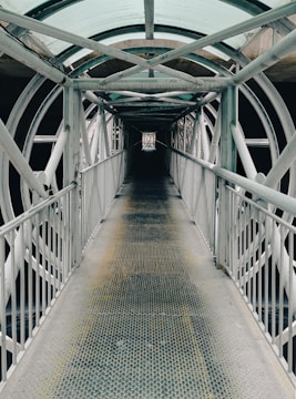 A long, narrow metal walkway extends forward, enclosed by a series of intersecting metal beams and a transparent, arched roof. The walkway is flanked by metal railings on both sides. The structure creates a tunnel-like effect with repeating patterns of geometric shapes, providing an industrial and symmetrical aesthetic.