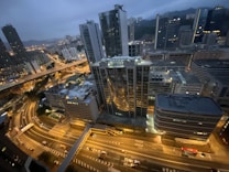 A nighttime cityscape featuring tall skyscrapers with illuminated windows. The image includes well-lit streets with vehicles and a complex network of roads and intersections. In the background, there are hills and more high-rise buildings, partially obscured by the evening haze.