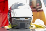 A carbon fiber racing helmet featuring a glossy finish and a reflective visor sits on a flat surface. The helmet bears the logo of 'Bell Helmets' in a small oval on the front. It is surrounded by soft cleaning cloths, and hints of a red background are visible.