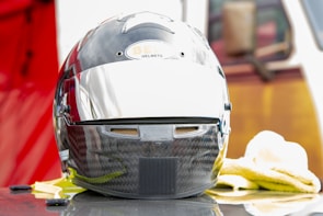 A carbon fiber racing helmet featuring a glossy finish and a reflective visor sits on a flat surface. The helmet bears the logo of 'Bell Helmets' in a small oval on the front. It is surrounded by soft cleaning cloths, and hints of a red background are visible.