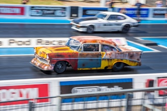 A vintage, rust-colored car is racing down a dragstrip alongside a white sports car. The scene depicts a high-speed race on a professional track with blurred motion indicating speed.