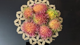 A group of vibrant, spiky rambutan fruits with a mix of pink, red, and green hues is arranged on an intricately designed woven pattern mat. The background is a solid black surface which contrasts with the bright colors of the fruits.