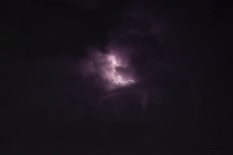 A dark sky is pierced by a flash of lightning, partially illuminating the surrounding clouds. The scene captures the intensity and power of a stormy night.