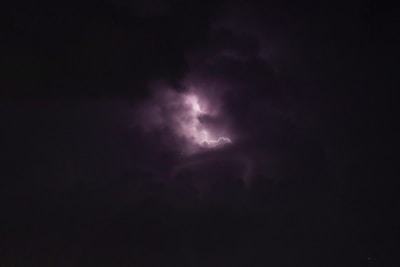 A dark sky is pierced by a flash of lightning, partially illuminating the surrounding clouds. The scene captures the intensity and power of a stormy night.
