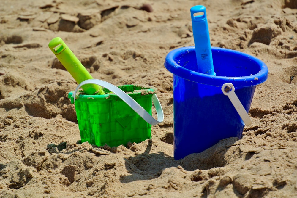 two plastic buckets with green straws in sand