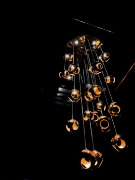 A modern chandelier with multiple spherical light fixtures hangs from the ceiling. Each fixture is metallic and polished, reflecting light in a warm, golden hue. The overall lighting creates a contrast against the dark background, making the chandelier the focal point of the image, emitting a cozy ambiance.