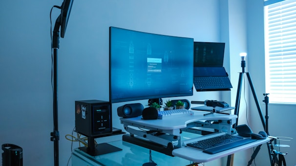 A modern workstation setup featuring a large curved monitor displaying a blue screen with digital diagrams. A laptop stand holds a second, inclined laptop. The desk is equipped with a speaker, microphone, keyboard, and a small plant for decoration. A soft light illuminates the area from a tall standing lamp near a window with blinds partially open.