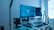 A modern workstation setup featuring a large curved monitor displaying a blue screen with digital diagrams. A laptop stand holds a second, inclined laptop. The desk is equipped with a speaker, microphone, keyboard, and a small plant for decoration. A soft light illuminates the area from a tall standing lamp near a window with blinds partially open.