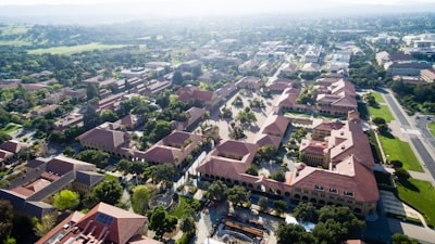 An aerial view of a university campus featuring numerous red-roofed buildings arranged in a grid pattern. The landscape includes a variety of trees and greenery, and pathways separating the buildings. In the background, the distant landscape fades into misty hills.