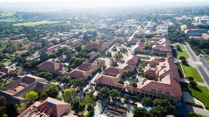 An aerial view of a university campus featuring numerous red-roofed buildings arranged in a grid pattern. The landscape includes a variety of trees and greenery, and pathways separating the buildings. In the background, the distant landscape fades into misty hills.