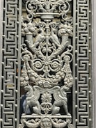 Intricately designed metalwork featuring symmetrical patterns with detailed carvings of mythological creatures, birds, and floral motifs. The central design consists of two birds facing each other perched on decorative cornucopias, surrounded by elaborate leaves and swirls. Lions' heads and griffin-like creatures enhance the ornate detailing, while the border has repetitive geometric patterns.