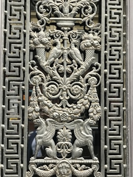 Intricately designed metalwork featuring symmetrical patterns with detailed carvings of mythological creatures, birds, and floral motifs. The central design consists of two birds facing each other perched on decorative cornucopias, surrounded by elaborate leaves and swirls. Lions' heads and griffin-like creatures enhance the ornate detailing, while the border has repetitive geometric patterns.