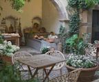 A charming outdoor courtyard with a rustic aesthetic, featuring a wooden table and chairs surrounded by lush greenery and potted flowers. The scene includes an arched stone wall covered with ivy and a rustic display of baskets filled with various vegetables and fruits, hinting at a market or garden supply area.