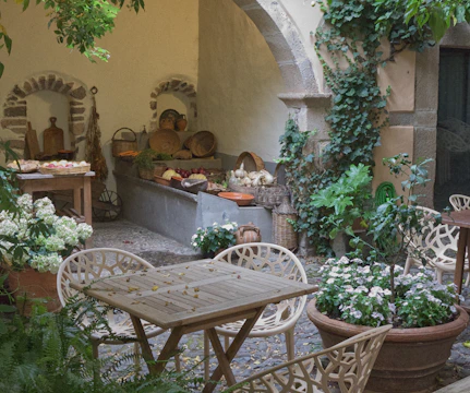 A charming outdoor courtyard with a rustic aesthetic, featuring a wooden table and chairs surrounded by lush greenery and potted flowers. The scene includes an arched stone wall covered with ivy and a rustic display of baskets filled with various vegetables and fruits, hinting at a market or garden supply area.