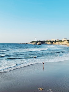 A scenic coastal view featuring a sandy beach with several people enjoying the water. The ocean waves gently lap against the shore, and in the distance, a lighthouse stands on a rocky outcrop. Buildings are perched on the cliffs, under a clear blue sky.
