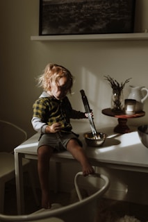 A small child with light brown hair sits on a white table, holding a pair of tongs and a mixing bowl. The child is wearing a plaid shirt and grey shorts, with sunlight gently illuminating their face and casting shadows. Behind them, there is a white ceramic pitcher, a candle, and dried plants arranged on a wooden stand. A black and white landscape photo hangs above a shelf in the background.