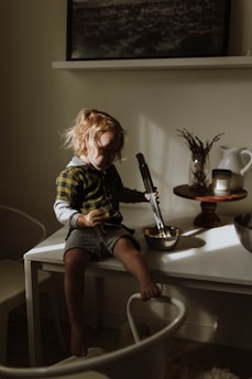 A small child with light brown hair sits on a white table, holding a pair of tongs and a mixing bowl. The child is wearing a plaid shirt and grey shorts, with sunlight gently illuminating their face and casting shadows. Behind them, there is a white ceramic pitcher, a candle, and dried plants arranged on a wooden stand. A black and white landscape photo hangs above a shelf in the background.