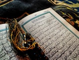 An open Arabic book with intricate calligraphy lying on a decorative fabric. A string of black prayer beads with tassels is placed across the pages, adding a contrast against the white and blue pages of the book.