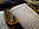 An open Arabic book with intricate calligraphy lying on a decorative fabric. A string of black prayer beads with tassels is placed across the pages, adding a contrast against the white and blue pages of the book.