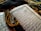 An open Arabic book with intricate calligraphy lying on a decorative fabric. A string of black prayer beads with tassels is placed across the pages, adding a contrast against the white and blue pages of the book.