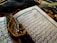 An open Arabic book with intricate calligraphy lying on a decorative fabric. A string of black prayer beads with tassels is placed across the pages, adding a contrast against the white and blue pages of the book.