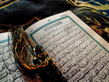 An open Arabic book with intricate calligraphy lying on a decorative fabric. A string of black prayer beads with tassels is placed across the pages, adding a contrast against the white and blue pages of the book.