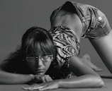 A person wearing glasses and a patterned top is performing a dynamic pose with their body bent and leaning forward on the floor. They are wearing denim shorts, and their expression is focused. The image is in black and white, highlighting shadows and contours.