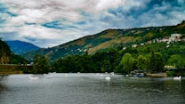 A serene river flows through a verdant landscape with rolling hills in the background. Small paddleboats are visible on the water, and lush green trees line the riverbanks. A few scattered houses are nestled on the hillside, and a sign reading 'Pirali' is seen on the right.