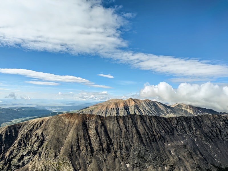 A broad brown mountain ridge rising above forested valleys under a blue sky with drifting white clouds