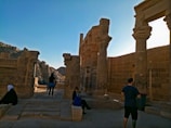 Ancient stone columns and partially ruins with people casually walking or sitting, creating a scene of exploration. The setting suggests historical significance, with a clear blue sky and rocky landscape surrounding the structures.