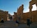 Ancient stone columns and partially ruins with people casually walking or sitting, creating a scene of exploration. The setting suggests historical significance, with a clear blue sky and rocky landscape surrounding the structures.