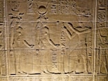 Ancient Egyptian wall carvings depicting figures in traditional attire and headgear, with hieroglyphics surrounding the scene. The figures appear to be engaged in some form of ritual or offering.