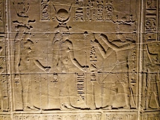 Ancient Egyptian wall carvings depicting figures in traditional attire and headgear, with hieroglyphics surrounding the scene. The figures appear to be engaged in some form of ritual or offering.