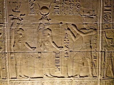 Ancient Egyptian wall carvings depicting figures in traditional attire and headgear, with hieroglyphics surrounding the scene. The figures appear to be engaged in some form of ritual or offering.