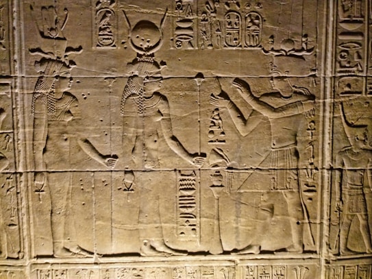 Ancient Egyptian wall carvings depicting figures in traditional attire and headgear, with hieroglyphics surrounding the scene. The figures appear to be engaged in some form of ritual or offering.