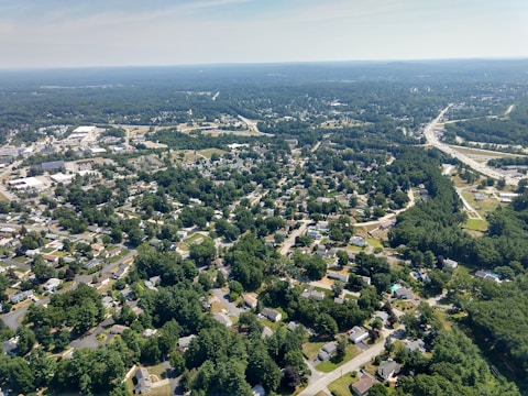 Aerial view of a suburban area with numerous houses, roads, and patches of dense trees. The landscape includes some commercial buildings and pathways, all set against a backdrop of vast greenery and open sky.
